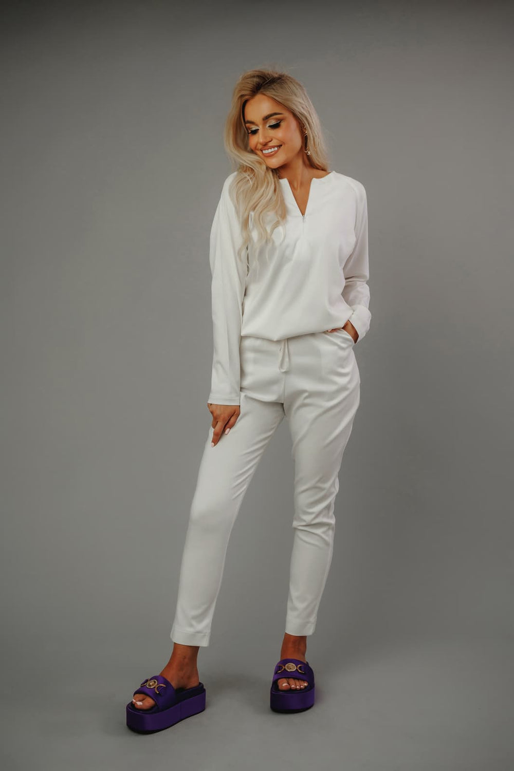 Uśmiechnięta_modelka_w_białej_bluzie_i_spodniach_MedElite_na_szarym_tle_fioletowe_designerskie_obuwie